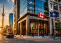 A city street at sunset with people walking and cars driving. Northstar Bank, a leader in Defence Security and Resilience, stands with Canadian and NATO flags outside. The CN Tower rises in the background as sunlight glows on buildings and street.
