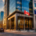 A city street at sunset with people walking and cars driving. Northstar Bank, a leader in Defence Security and Resilience, stands with Canadian and NATO flags outside. The CN Tower rises in the background as sunlight glows on buildings and street.