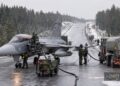 A grey Saab Gripen fighter jet refuels on a wintry, snow-dusted road surrounded by forest. Several crew members in green uniforms work around the aircraft, with hoses connected to fuel trucks among tall pine trees.
