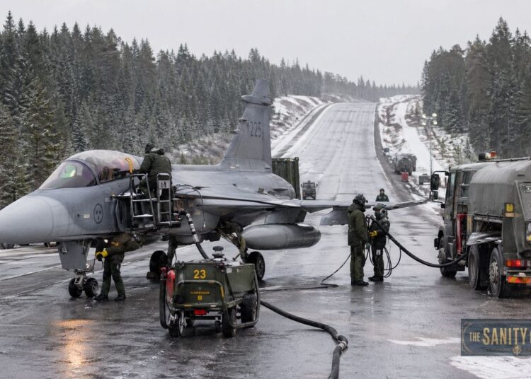 A grey Saab Gripen fighter jet refuels on a wintry, snow-dusted road surrounded by forest. Several crew members in green uniforms work around the aircraft, with hoses connected to fuel trucks among tall pine trees.