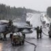 A grey Saab Gripen fighter jet refuels on a wintry, snow-dusted road surrounded by forest. Several crew members in green uniforms work around the aircraft, with hoses connected to fuel trucks among tall pine trees.