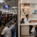 Split image: Left side shows a crowded, busy hospital waiting area with people lined up at a registration desk under “OPD” signs. Right side highlights healthcare privatization—a modern clinic where a doctor warmly consults with a patient in a bright office.