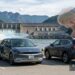Two cars are parked near a hydroelectric dam and EV charging station, illustrating electric vehicles vs gas cars. One car charges with a blue glow; the other is surrounded by smoke and an orange explosion. Mountains and water form the backdrop: “The Sanity Project.”.