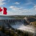 A large Canadian flag waves near a hydroelectric dam on the Canada Electricity Grid, with water flowing out. Wind turbines and nuclear power plants stand in the background amid hills, trees, power lines, and a partly cloudy sky.