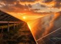Rows of solar panels reflect the golden light of a dramatic sunset, symbolizing the solar energy transition. The sky is partly cloudy, and sunlight illuminates the grassy field beneath, highlighting renewable technology in a peaceful, natural landscape.