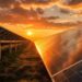 Rows of solar panels reflect the golden light of a dramatic sunset, symbolizing the solar energy transition. The sky is partly cloudy, and sunlight illuminates the grassy field beneath, highlighting renewable technology in a peaceful, natural landscape.