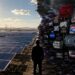 A person stands at the divide between a sunny field of solar panels and wind turbines on the left, and a dark, chaotic wall of glitching TV screens and protest signs spreading renewable energy misinformation on the right.