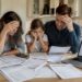 A worried family—mother, father, and daughter—sit at a kitchen table covered with bills, statements, and overdue notices. With canadian household debt rising, a laptop displays an “Interest Rates” chart trending upward.